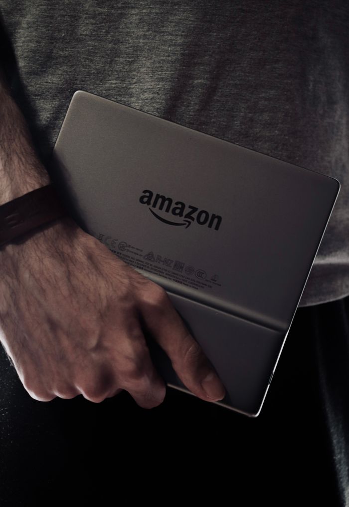 Close-up of a man's hand holding an Amazon Kindle device in a dimly lit setting.